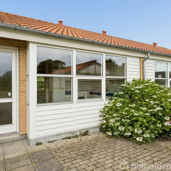 Rækkehus med rødt tegltag og store vinduer, flankeret af grønne buske og lyserøde blomster, beliggende i en brolagt forhave i et beboelsesområde.