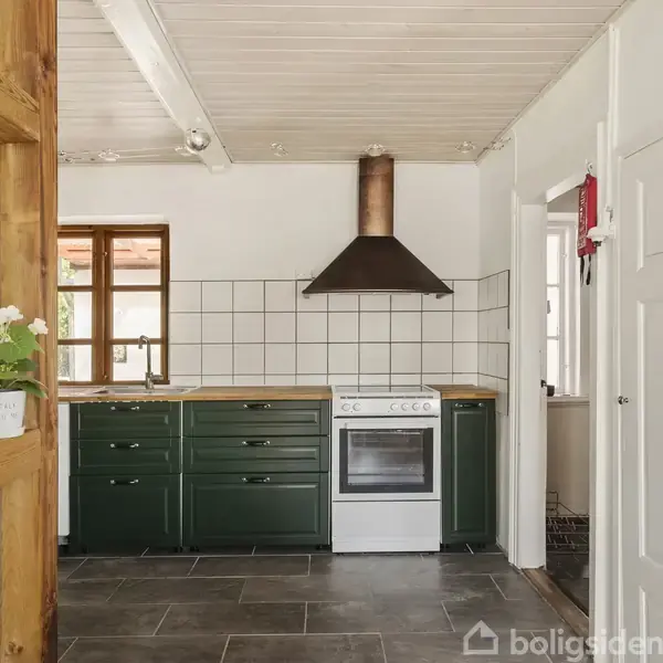 Et køkken med mørkegrønne skabe og en hvid ovn er i fokus. Ovnen har en mørk emhætte. Rummet har hvide vægge og loft samt en mørk flisegulvbelægning.