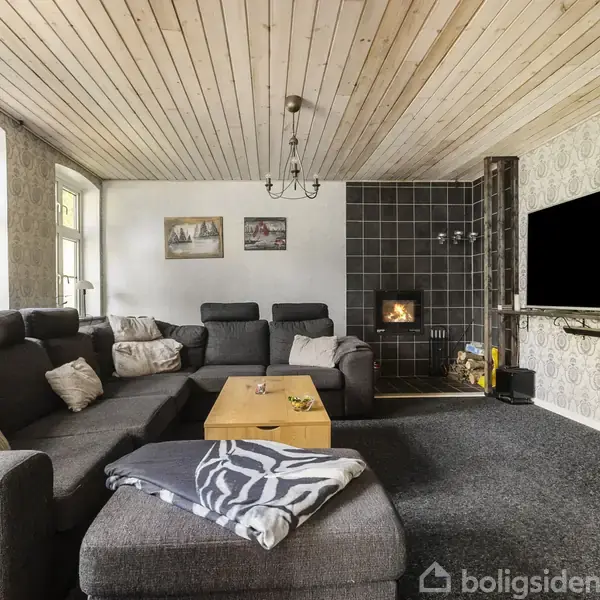 Stor, grå hjørnesofa med puder står foran et rektangulært sofabord. Pejsen er tændt ved væggen, og en stor fladskærms-tv hænger ved siden af. Rummet har en træbeklædt loft og et tæppebelagt gulv.