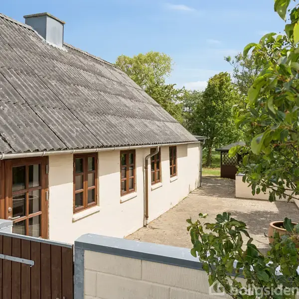 Et hvidt murstenshus med skråt tag og brune vinduer står på en gårdsplads omgivet af grønne træer. En lav mur med port adskiller gårdspladsen fra haven.