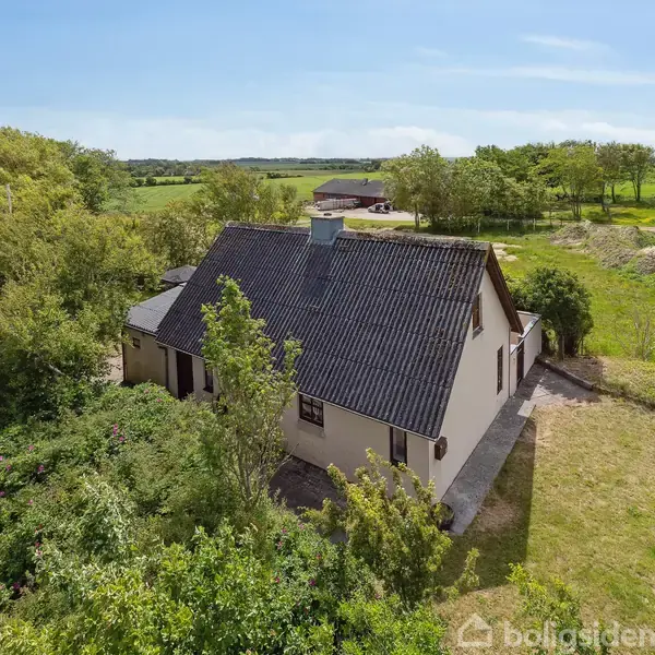 Et hvidt hus med sort tag omgivet af træer, står stille i et åbent naturlandskab med marker og småbygninger i baggrunden under en blå himmel.