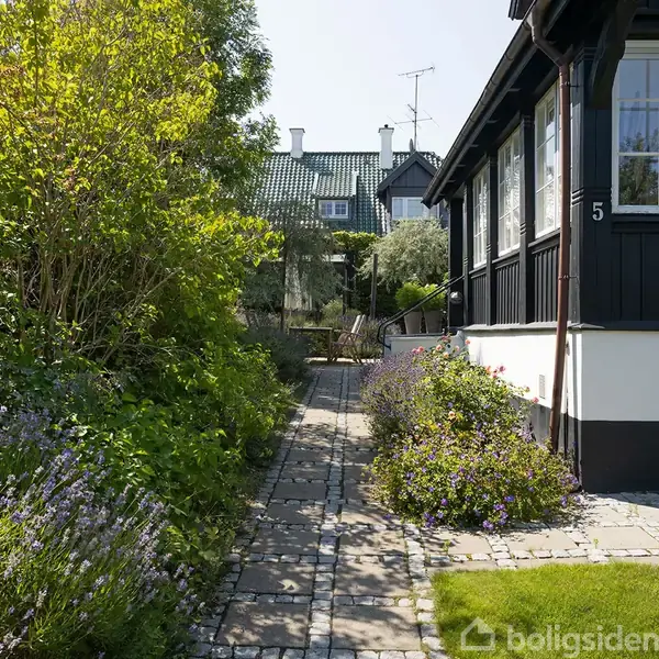 Et hus med sort træfacade og store vinduer ligger til højre. En smal flisebelagt sti fører gennem en frodig have med buske og blomster. Nummer "5" er synligt på huset.