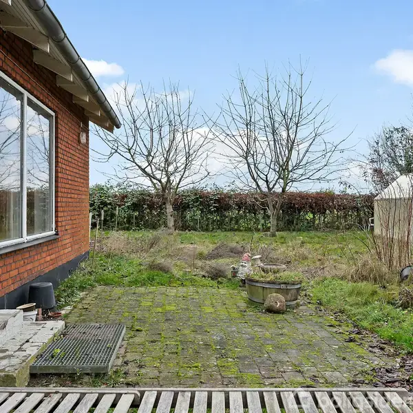 En terrasse af sten med en potteplante tæt på en rød murstensbygning. I baggrunden er der græsplæne, buske og træer samt et lille drivhus. Himmel med lette skyer.