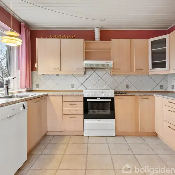 Et lyst køkken med træskabe og skuffer. Der er en ovn med emhætte, en opvaskemaskine til venstre og flisebelagt væg. Et vindue med gardiner tilføjer lys.