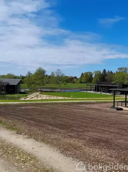 En åben plads med bare jorde i forgrunden, små bygninger i baggrunden, omgivet af grønne marker og træer, under en klar blå himmel.
