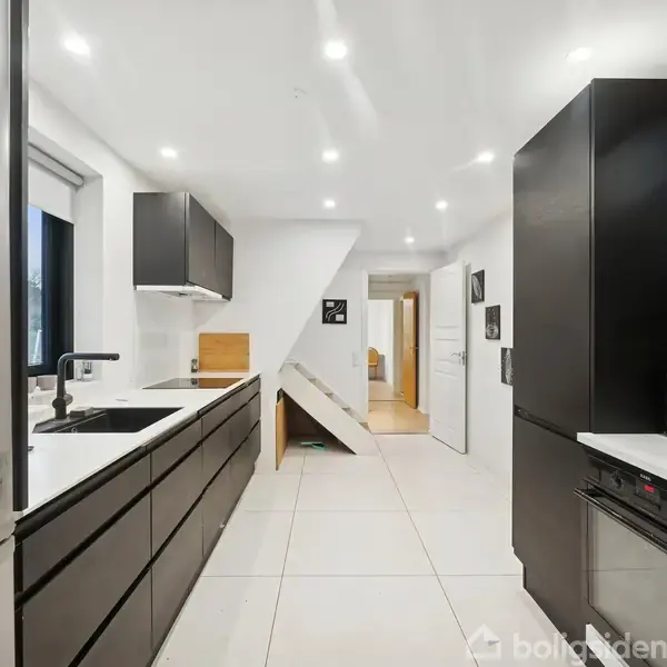 Et moderne køkken med sorte skabe og hvide bordplader, indeholder en stålvaske, kogeplader og diverse køkkenudstyr. Rummet har lyse fliser og indbyggede spots i loftet.