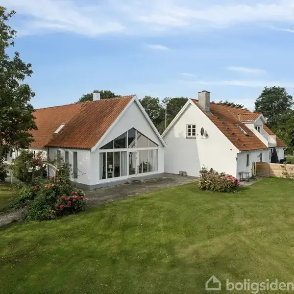 Et hvidt hus med rødt tegltag står stille i en grøn have med træer og buske omkring. Der er en stor terrasse foran huset.