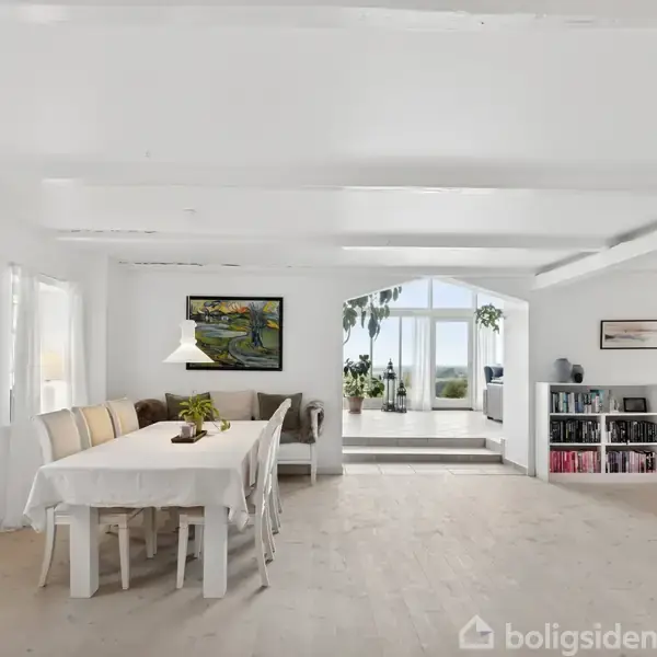 En lys stue med et spisebord med hvide stole, omgivet af malerier på væggene. En bogreol til højre, og glasdøre fører til en terrasse med udsigt til haven.