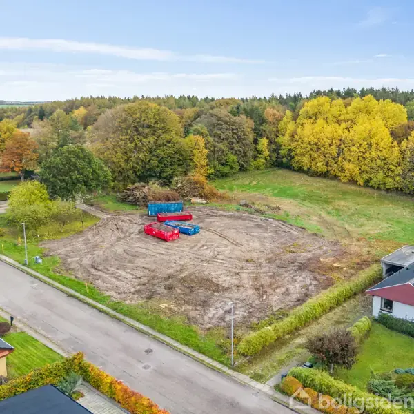 En jordet grund med flere farverige containere placeret centralt. Omkring ligger huse langs en lille vej, omgivet af grønne træer og marker under en klar blå himmel.