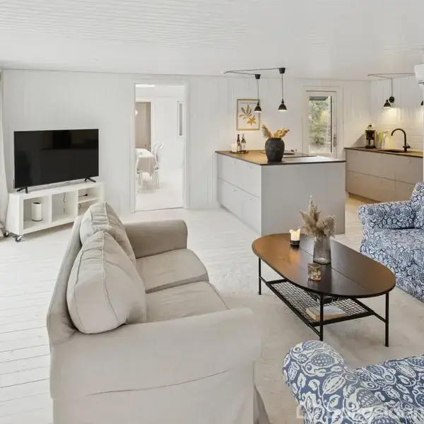 Stue med beige sofa og blåmønstret lænestol vender mod fladskærms-tv; køkken i baggrunden med grå ø og belysning, omgivet af store vinduer og hvidt interiør.