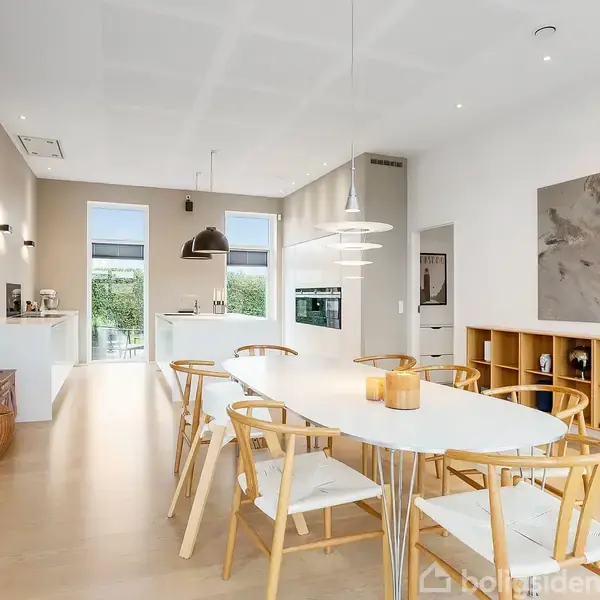 En stor, lys spisestue med et ovalt bord omgivet af træstole. Rummet har moderne indretning med kunst på væggen og åbner op mod et køkken med store vinduer.