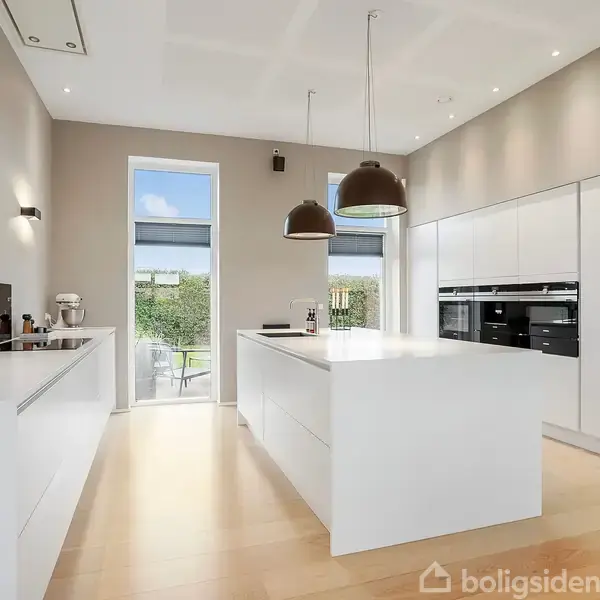 Et moderne, lyst køkken med en stor hvid køkkenø. To hængelamper hænger over øen. Rummet har indbyggede skabe og ovne, trægulv og store vinduer, der leder ud til en hæk.