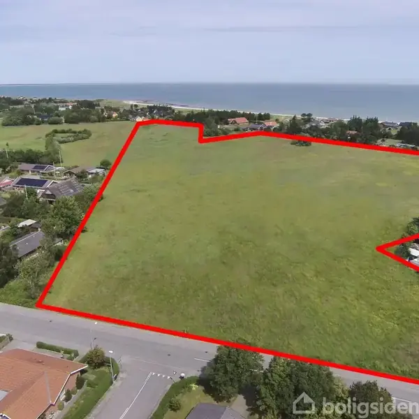 En stor, åben græsmark er indrammet af en rød linje, der angiver grunden. Den ligger i et boligområde nær havet, omgivet af huse og veje.