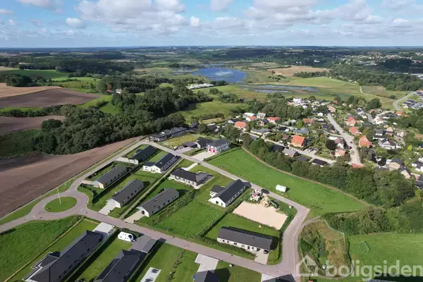 Landsby set fra luften viser rækkehuse og bygninger omgivet af marker og skov. En flod eller sø ses i baggrunden, mens veje forbinder området.