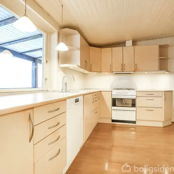 Lyst køkken med træskabe, hvidevarer og hvid bordplade. Køkkenet har ovn, opvaskemaskine og vask. Store vinduer slipper naturligt lys ind, mens træloftet og lamperne giver et varmt udtryk.