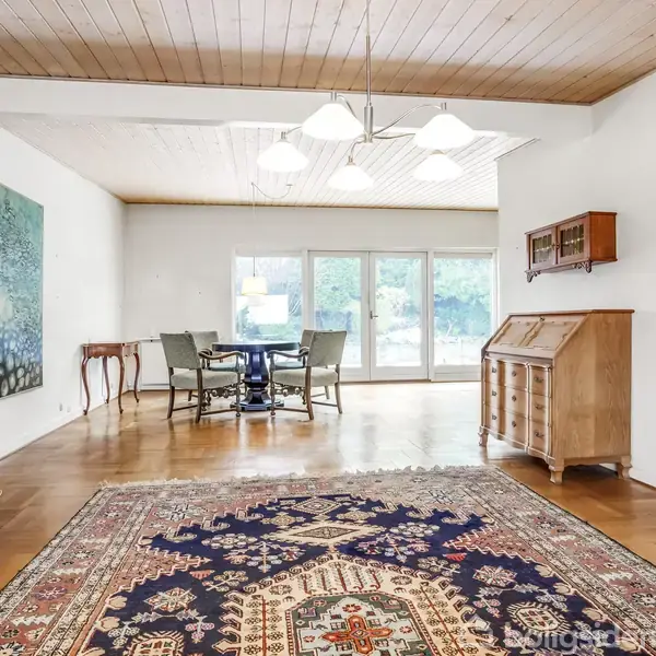 En stue med et stort, farverigt tæppe ligger på et trægulv. I baggrunden står et rundt spisebord med stole foran store vinduer. En skænk og kunst pryder væggene.