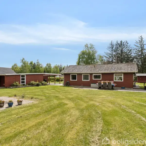 Et rødt hus står stille på en velplejet græsplæne med havemøbler foran. I baggrunden er der træer og en blå himmel.