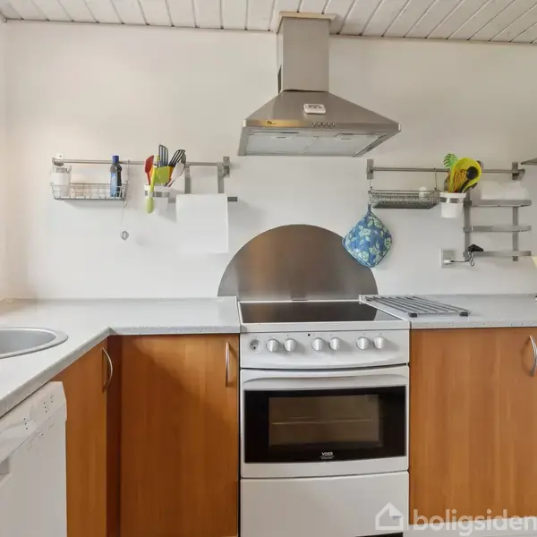 Komfur står i et kompakt køkken med hvide vægge og træskabe. På væggen er der hylder med køkkenredskaber og en kedel står på bordpladen ved siden af vasken.