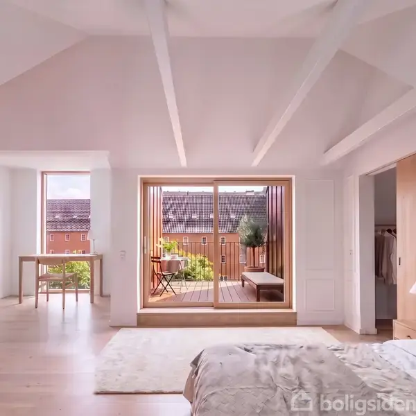 Soveværelse med seng, skrivebord og skydedøre åbner til en altan med udsigt over tage. Lyse træbjælker og gulve skaber en lys atmosfære. Tekst: Visualisering.