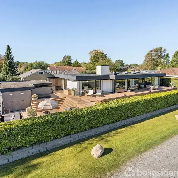 Et moderne enfamiliehus med fladt tag står på en velholdt grund omgivet af hække. Foran huset findes en stor træterrasse med siddepladser. Området er et roligt boligkvarter.
