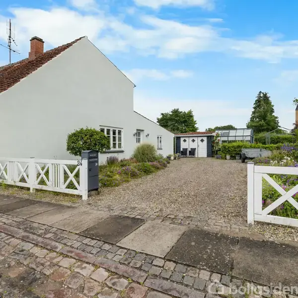 Et lyst køkken med hvide skabe og sorte fliser fører til en stue med trappe. Et spisebord med planter ved vinduet. Lamper hænger fra loftet.