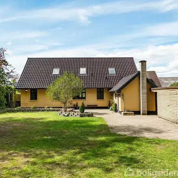 Et gult hus med mørkt tag står på en græsplæne. I baghaven ses en træterrasse og et lille skur. Himlen er blå med spredte skyer.