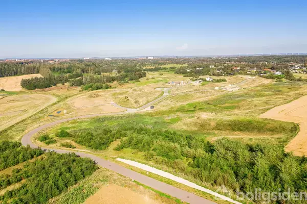 Grønne bakker strækker sig gennem et stort åbent landskab med spredt bebyggelse i baggrunden. En snoet vej skærer gennem området under en klar blå himmel.