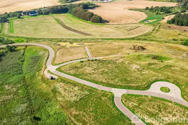 En snoet vej bevæger sig gennem et åbent, grønt landskab med marker og et par bygninger i baggrunden. Bakker og beplantning omgiver området.