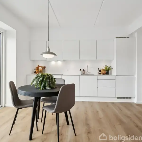 Et rundt sort spisebord med fire stole står i et lyst køkken med hvide skabe. Der er en hængende lampe over bordet og en glaspaneldør til venstre.