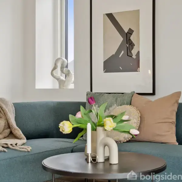 En blå sofa er dekoreret med puder og tæpper. På et rundt bord foran står en vase med tulipaner. Billedramme på væggen. Skulptur i vindueskarmen.
