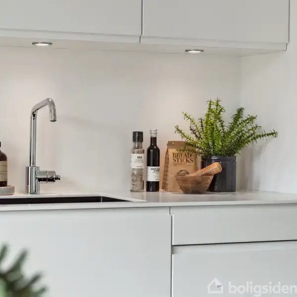 En køkkenvask med hane står i en modernistisk hvid køkkenindretning. Ved siden af vasken er der en brun sæbedispenser, plante, oliemasker og plastikflasker. Baggrunden er et hvidt skab.