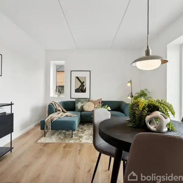En moderne stue med grøn sofa placeret ved et stort vindue. Et spisebord står på trægulvet med planter som dekoration. Væggene er hvide med minimalistisk kunst.