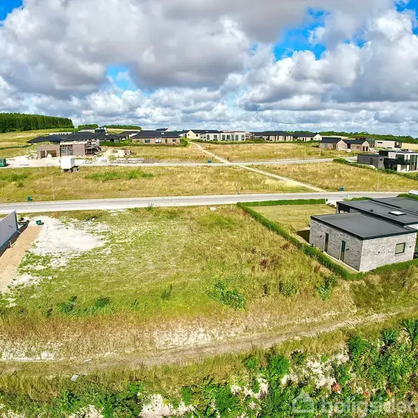 Moderne hus under opførelse står på en grund omgivet af grønt landskab. Sollys skinner gennem skyerne. Flere huse ses i baggrunden langs en vej.