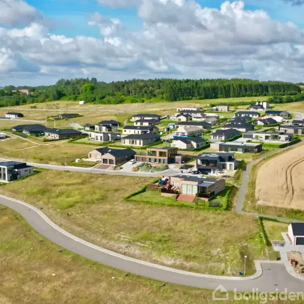 Boliger ligger spredt i en nyopført forstad i kuperet landskab. Veje forbinder husene, og marker omkranser området. Træer og skyer i baggrunden understreger den landlige stemning.