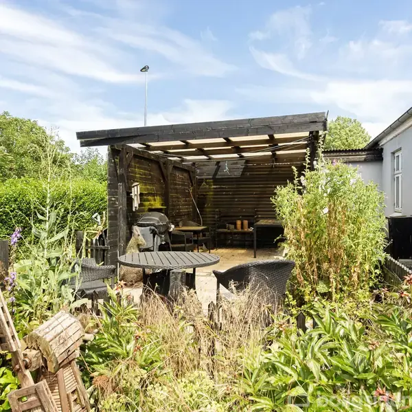 Overdækket terrasse med havemøbler står tæt ved et hvidt hus. Terrassebordet er omgivet af frodige planter og blomster, mens hækken i baggrunden indrammer haven.