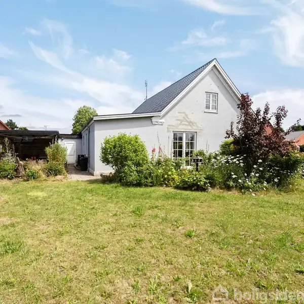 Hvidt hus med skråt tag står stille i en grøn have med blomster og buske; en garage ses i baggrunden under en blå himmel.