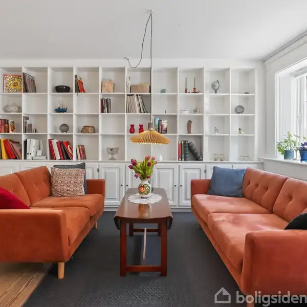 To orange sofaer står overfor hinanden med et lille bord imellem, omgivet af en stor, hvid reol fyldt med bøger og nipsting. Store vinduer giver naturligt lys.