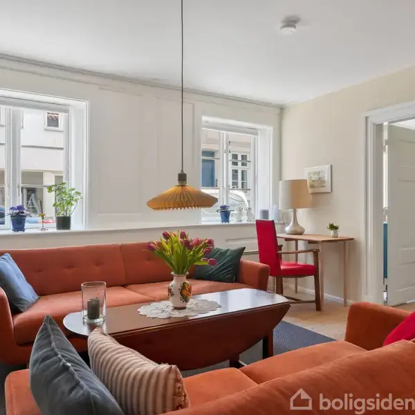 Stue med orange sofa omkring et sofabord. En vase med blomster står på bordet. Tre vinduer bagved med planter. Dør åbner til et andet rum med en seng.