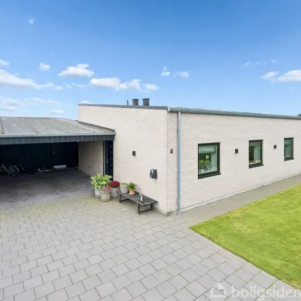 En moderne, hvid murstensbygning ses med en carport til venstre. Bygningen har sorte vinduer, og der er en velholdt græsplæne til højre. Himmelen er blå med få skyer.