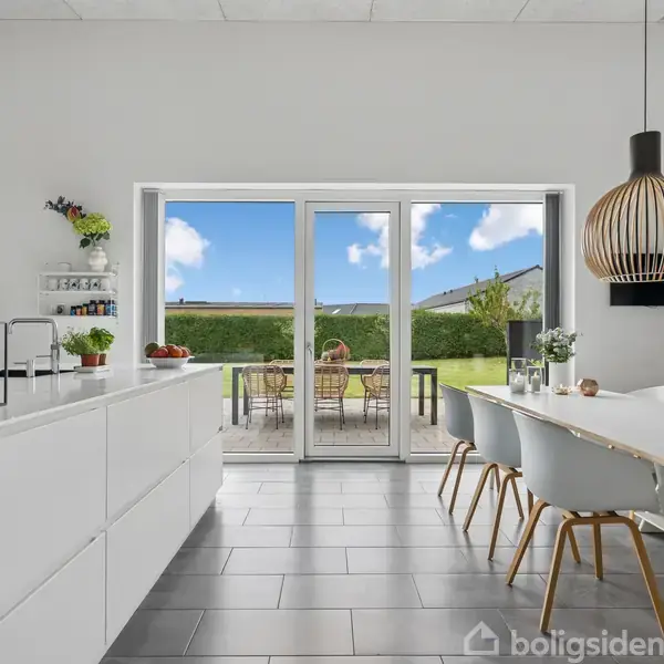 Et lyst køkken med hvide skabe og en træspisebord. Stole står ved bordet under en stor lampe. Store vinduer åbner til en have med græs og blå himmel.