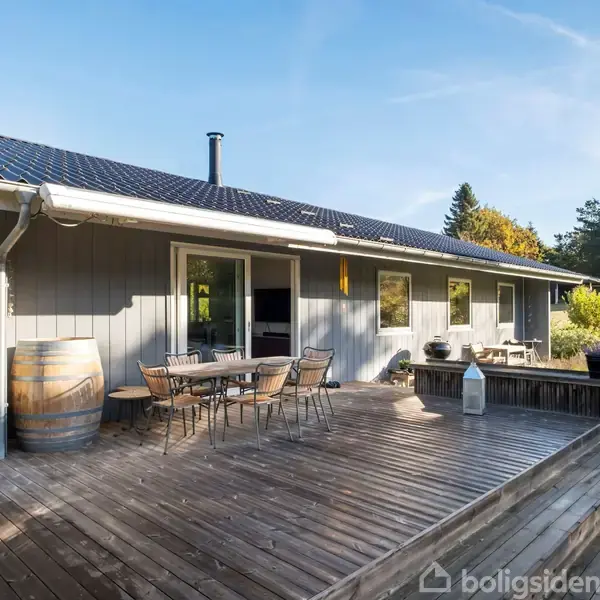 Et træhus har en terrasse med havemøbler. En vintønde står ved siden af bordet. I baggrunden er der en have med græs og nogle træer under en klar blå himmel.