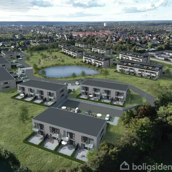 Rækkehuse med flade tage er placeret ved en kunstig sø, omgivet af grønt græs og skov. Flere husblokke ses i baggrunden med en by i horisonten.
