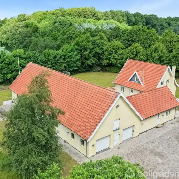 Et stort hus med gul facade og rødt tegltag ligger omgivet af frodig skov og åbne marker. Indkørslen er grusbelagt, og en græsplæne omgiver ejendommen.