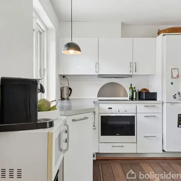 Et moderne køkken med hvide skabe og apparater. Køleskabet har magneter, og der står flasker på bordet. En lampe hænger ned fra loftet. Trægulve og lyse vægge omgiver rummet.