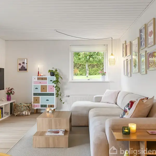 En lys stue med en beige hjørnesofa står foran en væg med farverige billeder. Et tv er monteret på væggen til venstre. Vindue viser grønt landskab udenfor.