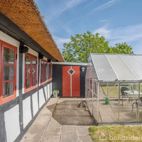 Bindingsværkshus med stråtag og røde vinduer står ved siden af et glasdrivhus. I baggrunden ses en græsplæne og en grøn busk under en blå himmel.