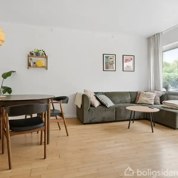 Sofahjørne med grå sofa og runde borde. Ved siden af er der et spisebord med stole. Store vinduer giver lys ind i den minimalistiske stue med træparketgulv og planter.