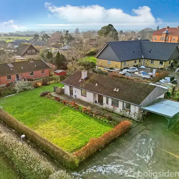Hus omgivet af en velplejet græsplæne med hække. I baggrunden er der flere andre huse og en gårdsplads med biler. Himlen er delvist overskyet.