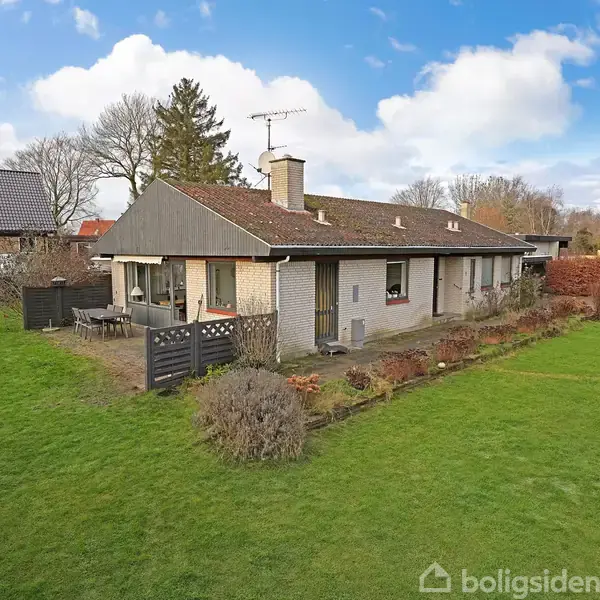 En lav, langstrakt villa med fladt tag står stille omgivet af en grøn have. Der er en lille terrasse med havemøbler, og huset omgives af velholdte buske.