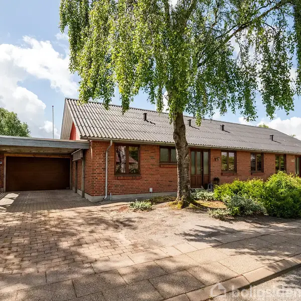 En rød murstensbungalow med garage indkørsel ved siden af, omgivet af grønne buske og et stort træ foran. Det er solskin, med nogle spredte skyer på himlen.